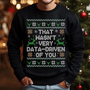 Pode incluir: Camisola preta de gola redonda com um design festivo. A camisola apresenta um padrão repetido de renas verdes e flocos de neve brancos. O texto "THAT WASN'T VERY DATA-DRIVEN OF YOU" é exibido em branco.