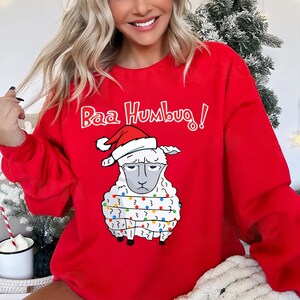 May include: Red sweatshirt with the text "Baa Humbug!" above a sheep wearing a Santa hat and Christmas lights. The sheep has a grumpy expression. A festive holiday-themed apparel item.