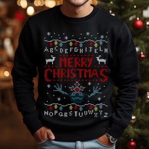 May include: Black crew neck sweatshirt with a festive Christmas design. The design includes the words "MERRY CHRISTMAS" in red, a string of colourful lights, snowflakes, and a stylized illustration. The letters of the alphabet are also included.