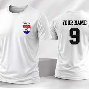 K&ouml;nnte beinhalten: Ein wei&szlig;es Kurzarm-T-Shirt mit dem kroatischen Wappen und dem Wort CROATIA auf der linken Brustseite. Die R&uuml;ckseite des T-Shirts zeigt den Text YOUR NAME in schwarzen Blockbuchstaben &uuml;ber der gro&szlig;en schwarzen Ziffer 9.