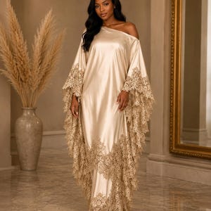 May include: A floor-length champagne-colored satin kaftan dress featuring an asymmetrical one-shoulder neckline and wide, flowing sleeves. The garment is adorned with intricate, scalloped beige lace trim along the sleeve edges and hemline.