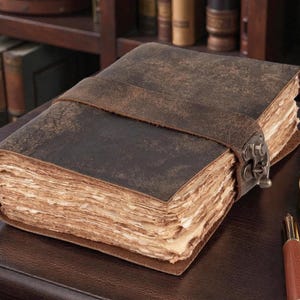 May include: A vintage-style leather journal with a distressed brown cover and a leather strap closure. The journal's pages have a rustic, aged appearance. A compass and pen are also visible.
