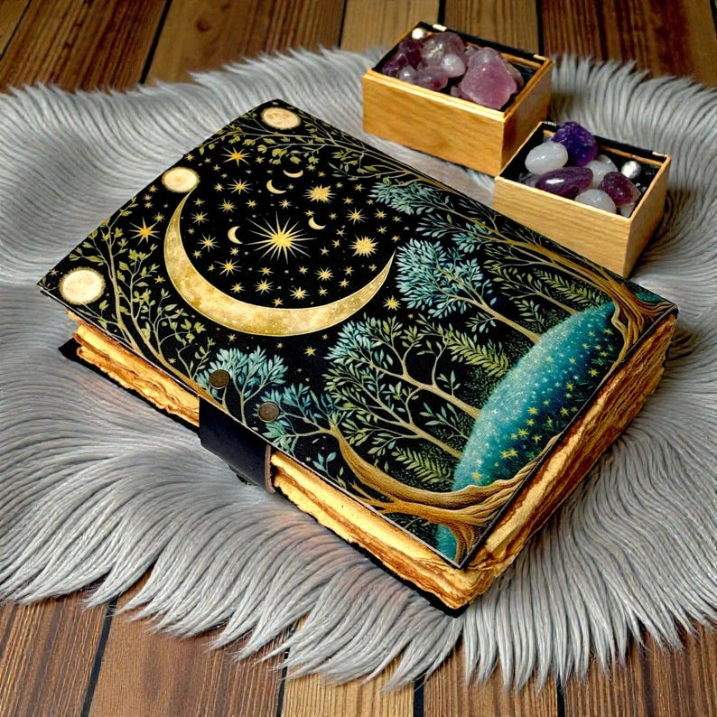 Puede incluir: Un diario decorativo con una cubierta negra con un dise&ntilde;o de luna creciente dorada, estrellas y &aacute;rboles. Las p&aacute;ginas del diario tienen un borde dorado. Dos peque&ntilde;as cajas de madera llenas de piedras preciosas est&aacute;n cerca.