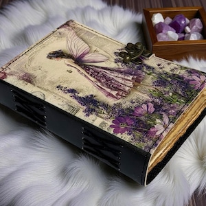 May include: A decorative journal with a fairy illustration on the cover, featuring a purple dress and lavender flowers. The journal has a black spine and a metal clasp. The pages have a vintage, aged appearance.