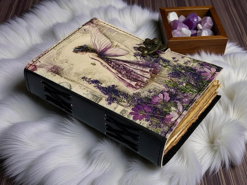 May include: A decorative journal with a fairy illustration on the cover, featuring a purple dress and lavender flowers. The journal has a black spine and a metal clasp. The pages have a vintage, aged appearance.