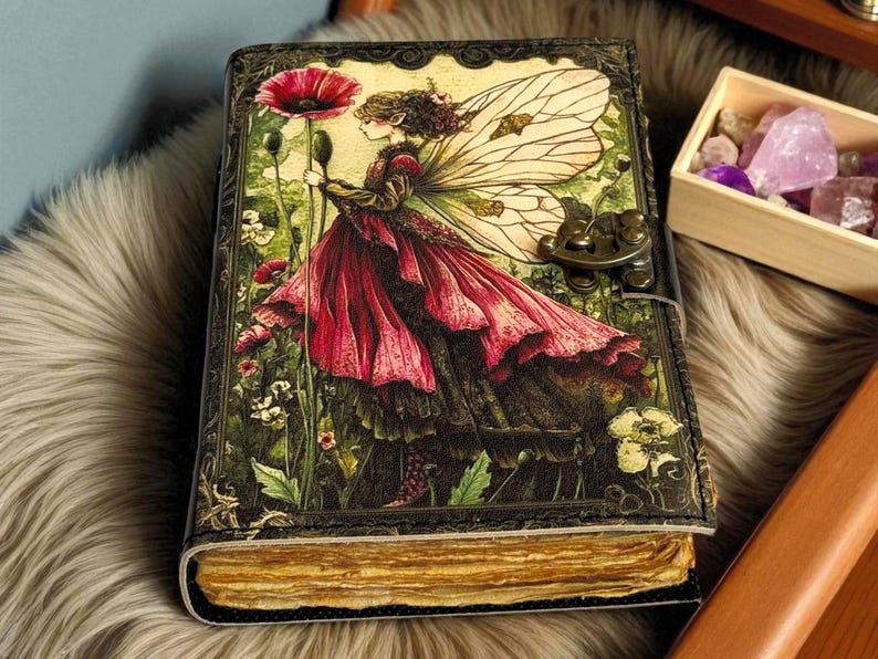 K&ouml;nnte beinhalten: Ein ledergebundenes Notizbuch mit einer Fantasy-Illustration. Das Cover zeigt eine Fee in einem roten Kleid, die eine Blume h&auml;lt, vor einem Hintergrund aus Blumen und Bl&auml;ttern. Das Notizbuch hat einen Metallverschluss und gealtertes Papier.