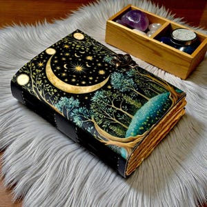 Puede incluir: Un diario de cuero negro con un dise&ntilde;o celestial que presenta una luna creciente, estrellas y &aacute;rboles. El diario tiene un cierre de metal y p&aacute;ginas envejecidas. Una caja de madera con cristales est&aacute; cerca.