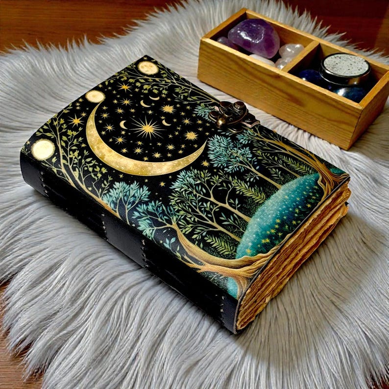 Puede incluir: Un diario de cuero negro con un dise&ntilde;o celestial que presenta una luna creciente, estrellas y &aacute;rboles. El diario tiene un cierre de metal y p&aacute;ginas envejecidas. Una caja de madera con cristales est&aacute; cerca.