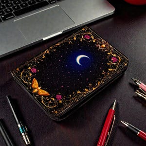 May include: A black journal with a celestial design featuring a crescent moon, stars, and a butterfly. The cover is adorned with gold floral accents and pink flowers. Pens and a laptop are in the background.