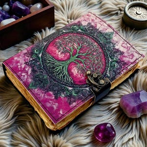Puede incluir: Un diario de cuero con una cubierta rosa y morada que presenta un dise&ntilde;o de &aacute;rbol de la vida verde. El diario tiene un cierre de metal y p&aacute;ginas con bordes dorados. Cristales de amatista y una caja de madera con gemas est&aacute;n cerca.