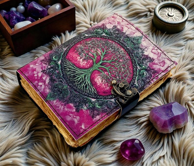Puede incluir: Un diario de cuero con una cubierta rosa y morada que presenta un dise&ntilde;o de &aacute;rbol de la vida verde. El diario tiene un cierre de metal y p&aacute;ginas con bordes dorados. Cristales de amatista y una caja de madera con gemas est&aacute;n cerca.