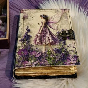 May include: A decorative journal with a fairy design. The cover features a fairy in a purple dress amidst purple flowers. The journal has a metal clasp and aged-looking pages. The overall theme is fantasy and nature.
