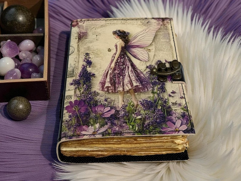 May include: A decorative journal with a fairy design. The cover features a fairy in a purple dress amidst purple flowers. The journal has a metal clasp and aged-looking pages. The overall theme is fantasy and nature.