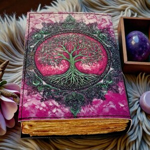 Puede incluir: Un diario hecho a mano con un dise&ntilde;o de &aacute;rbol de la vida rosa y verde en la portada. El diario tiene una cubierta de cuero y p&aacute;ginas de papel envejecido. Una gema p&uacute;rpura y una peque&ntilde;a caja de madera est&aacute;n junto al diario.