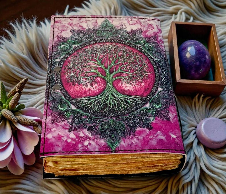 Puede incluir: Un diario hecho a mano con un dise&ntilde;o de &aacute;rbol de la vida rosa y verde en la portada. El diario tiene una cubierta de cuero y p&aacute;ginas de papel envejecido. Una gema p&uacute;rpura y una peque&ntilde;a caja de madera est&aacute;n junto al diario.
