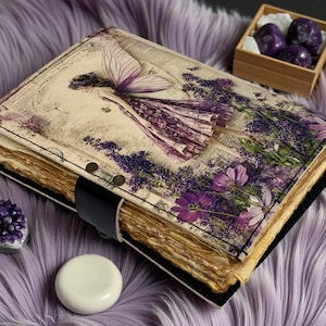 May include: A handmade journal with a fairy illustration on the cover, featuring a purple and lavender color scheme. The journal has a black leather strap closure and aged paper pages. Additional items include amethyst crystals and a wooden box.
