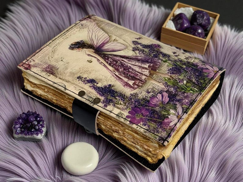 May include: A handmade journal with a fairy illustration on the cover, featuring a purple and lavender color scheme. The journal has a black leather strap closure and aged paper pages. Additional items include amethyst crystals and a wooden box.