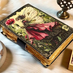 K&ouml;nnte beinhalten: Ein Tagebuch im Vintage-Stil mit einer Feenillustration auf dem Einband. Die Fee tr&auml;gt ein rotes Kleid und h&auml;lt eine rote Blume. Das Tagebuch hat gealterte Seiten und einen schwarzen Lederriemen.
