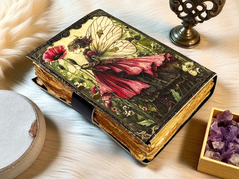 K&ouml;nnte beinhalten: Ein Tagebuch im Vintage-Stil mit einer Feenillustration auf dem Einband. Die Fee tr&auml;gt ein rotes Kleid und h&auml;lt eine rote Blume. Das Tagebuch hat gealterte Seiten und einen schwarzen Lederriemen.