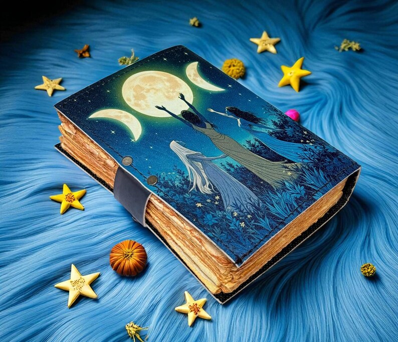 Puede incluir: Un diario decorativo con un dise&ntilde;o celestial que presenta una luna llena, lunas crecientes y figuras con vestidos fluidos. La cubierta es azul con una correa de cuero negro, y las p&aacute;ginas tienen una apariencia vintage envejecida. Decoraciones amarillas en forma de estrella rodean el diario.