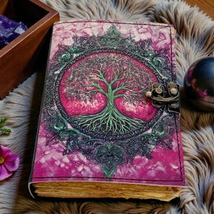 Puede incluir: Un diario de cuero con una cubierta rosa y morada que presenta un dise&ntilde;o detallado de &aacute;rbol de la vida. El diario tiene un cierre de metal y p&aacute;ginas envejecidas. Una caja de madera con cristales morados y una esfera de metal est&aacute;n cerca.
