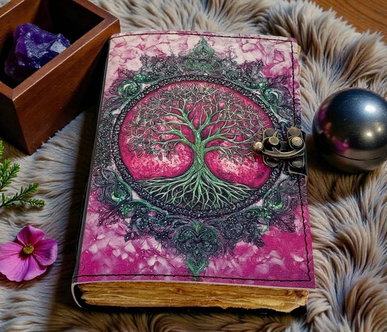 Puede incluir: Un diario de cuero con una cubierta rosa y morada que presenta un dise&ntilde;o detallado de &aacute;rbol de la vida. El diario tiene un cierre de metal y p&aacute;ginas envejecidas. Una caja de madera con cristales morados y una esfera de metal est&aacute;n cerca.