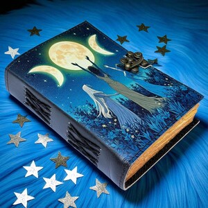 Puede incluir: Un diario encuadernado en cuero con un dise&ntilde;o celestial. La portada presenta una luna llena, lunas crecientes y figuras que se alzan hacia arriba sobre un fondo azul estrellado. El diario tiene un cierre de metal y encuadernaci&oacute;n cosida, con bordes de papel dorados.