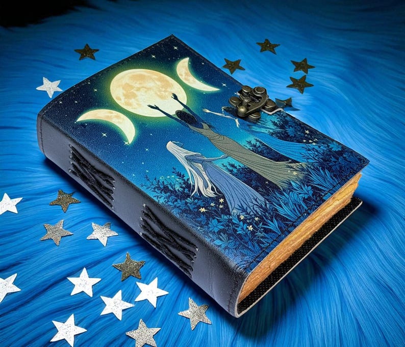 Puede incluir: Un diario encuadernado en cuero con un dise&ntilde;o celestial. La portada presenta una luna llena, lunas crecientes y figuras que se alzan hacia arriba sobre un fondo azul estrellado. El diario tiene un cierre de metal y encuadernaci&oacute;n cosida, con bordes de papel dorados.
