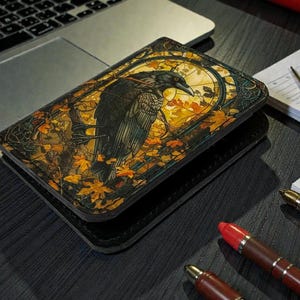 May include: A black leather wallet featuring a detailed illustration of a raven perched on a branch, set against a stained-glass style background with autumn leaves. The wallet is rectangular and appears to be closed.