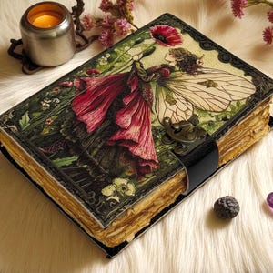 K&ouml;nnte beinhalten: Ein Vintage-Journal mit einer detaillierten Illustration einer Fee, die eine rote Blume h&auml;lt. Das Journal hat einen schwarzen Lederriemenverschluss und gealterte Papierseiten. Das Cover zeigt eine Fee mit gro&szlig;en Fl&uuml;geln und einem roten Kleid.