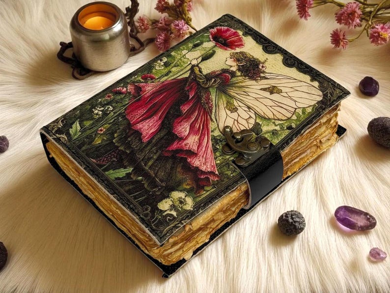 K&ouml;nnte beinhalten: Ein Vintage-Journal mit einer detaillierten Illustration einer Fee, die eine rote Blume h&auml;lt. Das Journal hat einen schwarzen Lederriemenverschluss und gealterte Papierseiten. Das Cover zeigt eine Fee mit gro&szlig;en Fl&uuml;geln und einem roten Kleid.