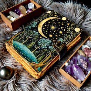 Puede incluir: Un diario decorativo con un dise&ntilde;o celestial que presenta una luna creciente, estrellas y &aacute;rboles. El diario tiene un cierre de correa de cuero y p&aacute;ginas de papel envejecido. Cajas de madera llenas de cristales y piedras preciosas tambi&eacute;n est&aacute;n en la imagen.