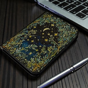 May include: A black notebook with a celestial design featuring a crescent moon, stars, and foliage in shades of green, blue, and gold. A silver pen rests beside the notebook on a dark wooden surface, near a laptop.