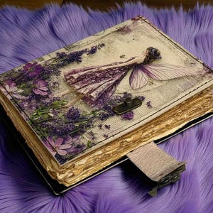 May include: A decorative journal with a fairy and floral design. The cover features a fairy illustration with purple and white accents, surrounded by lavender and flowers. The journal has a clasp closure and aged paper pages.