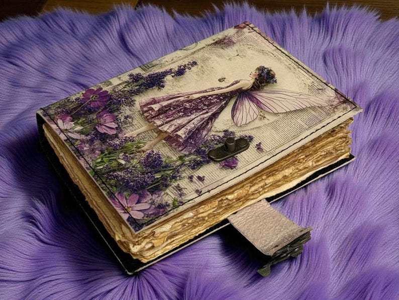 May include: A decorative journal with a fairy and floral design. The cover features a fairy illustration with purple and white accents, surrounded by lavender and flowers. The journal has a clasp closure and aged paper pages.