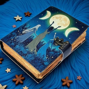 Puede incluir: Un diario decorativo con un dise&ntilde;o celestial que presenta una luna llena, lunas crecientes y figuras con vestidos fluidos. La cubierta es azul con un lomo negro y un cierre de metal. Estrellas y flores de madera rodean el diario sobre una superficie azul.