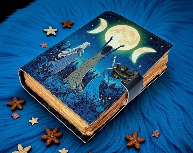 Puede incluir: Un diario decorativo con un dise&ntilde;o celestial que presenta una luna llena, lunas crecientes y figuras con vestidos fluidos. La cubierta es azul con un lomo negro y un cierre de metal. Estrellas y flores de madera rodean el diario sobre una superficie azul.