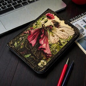 May include: A black wallet featuring a colorful illustration of a fairy holding a red flower. The fairy is wearing a red dress with large, detailed wings. The wallet is on a dark wooden surface near a laptop and pens.