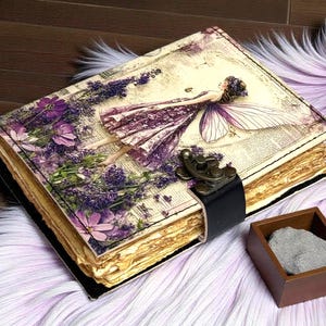 May include: A rectangular journal with a vintage-style cover featuring a fairy illustration, purple lavender flowers, and text. The book has aged, deckled-edge pages, a black leather strap closure with a metal latch, and sits beside a small wooden box.