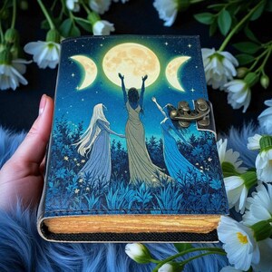 Puede incluir: Un diario decorativo con un dise&ntilde;o celestial. La portada presenta una luna llena y lunas crecientes, con tres figuras con vestidos fluidos. El diario tiene un cierre de lat&oacute;n y un borde texturizado de aspecto envejecido. La paleta de colores incluye azules, verdes y dorados.