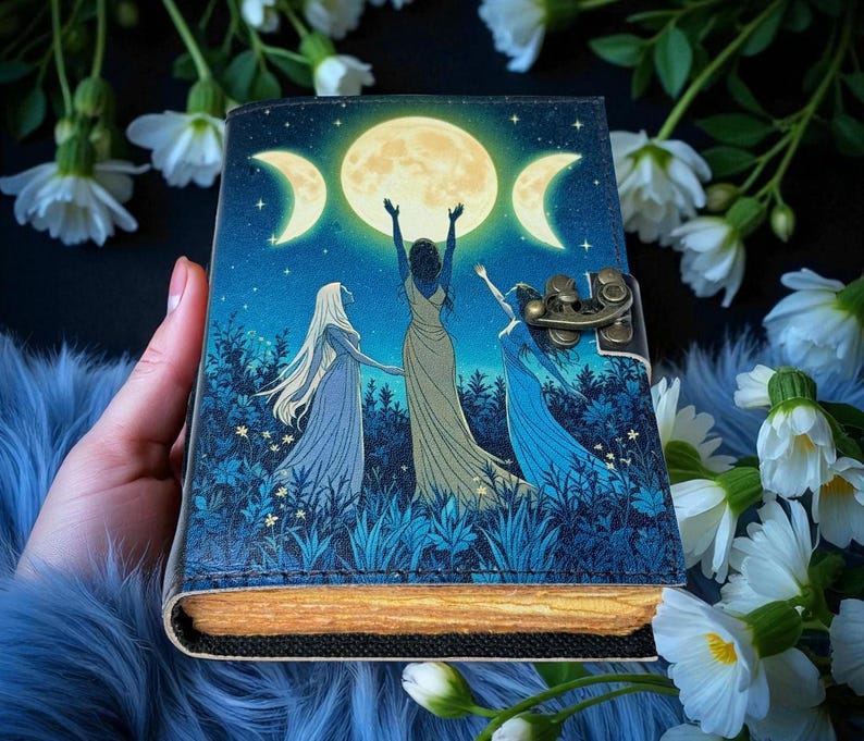 Puede incluir: Un diario decorativo con un dise&ntilde;o celestial. La portada presenta una luna llena y lunas crecientes, con tres figuras con vestidos fluidos. El diario tiene un cierre de lat&oacute;n y un borde texturizado de aspecto envejecido. La paleta de colores incluye azules, verdes y dorados.