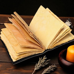 Puede incluir: Un diario abierto de estilo antiguo, con p&aacute;ginas amarillentas y envejecidas. Se ve un peque&ntilde;o marcap&aacute;ginas decorativo. Una vela naranja encendida en un portavelas de madera est&aacute; cerca, con ramitas de plantas secas en primer plano. El diario est&aacute; sobre una superficie de madera oscura.