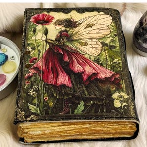 K&ouml;nnte beinhalten: Ein Tagebuch im Vintage-Stil mit einer Feenillustration auf dem Einband. Die Fee tr&auml;gt ein flie&szlig;endes rotes Kleid und h&auml;lt eine gro&szlig;e rote Blume. Der Einband hat einen dunklen, verzierten Rand, und die Seiten haben einen goldenen Farbton. Das Tagebuch ist mit einem schwarzen Riemen verschlossen.