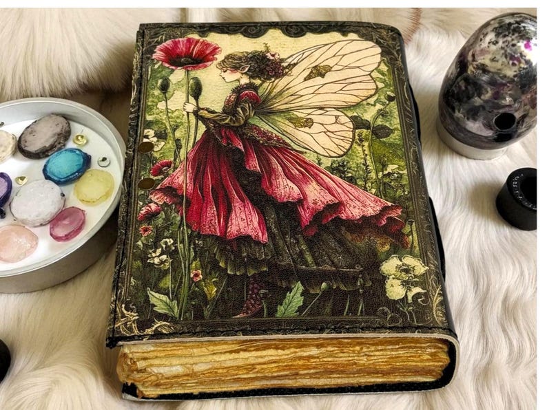 K&ouml;nnte beinhalten: Ein Tagebuch im Vintage-Stil mit einer Feenillustration auf dem Einband. Die Fee tr&auml;gt ein flie&szlig;endes rotes Kleid und h&auml;lt eine gro&szlig;e rote Blume. Der Einband hat einen dunklen, verzierten Rand, und die Seiten haben einen goldenen Farbton. Das Tagebuch ist mit einem schwarzen Riemen verschlossen.