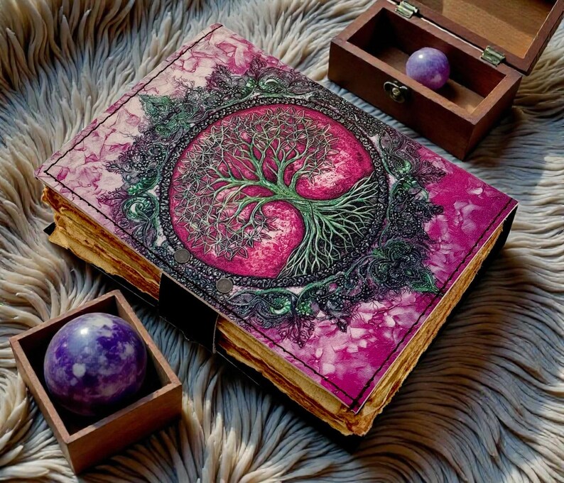 Puede incluir: Un diario hecho a mano con una cubierta rosa y morada con un dise&ntilde;o de &aacute;rbol de la vida. El diario tiene una encuadernaci&oacute;n de cuero y papel envejecido. Dos peque&ntilde;as cajas de madera contienen cada una una esfera p&uacute;rpura.