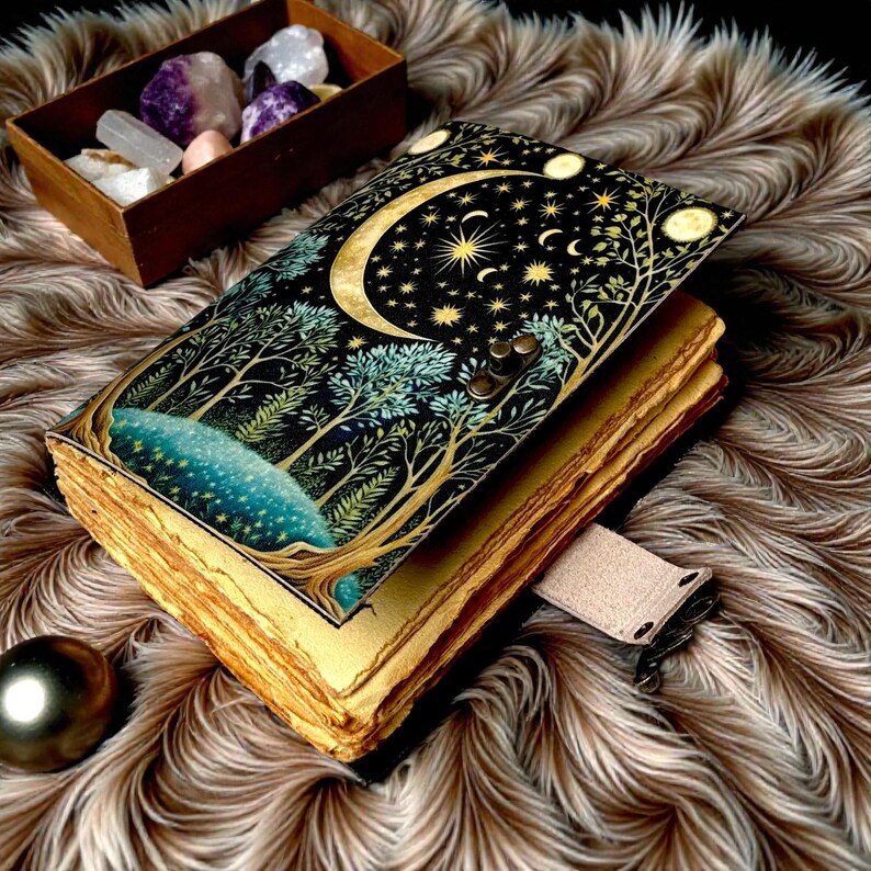 Puede incluir: Un diario decorativo con un dise&ntilde;o celestial que presenta una luna creciente, estrellas y &aacute;rboles. La cubierta es negra con detalles dorados y turquesas. Las p&aacute;ginas envejecidas son visibles y tiene una correa de cuero.