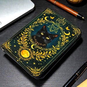 May include: A dark green wallet featuring a black cat with yellow eyes, surrounded by gold crescent moons, stars, and floral designs. The wallet has a celestial and mystical theme, with a pentagram and a full moon illustration.