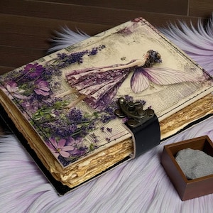 May include: A vintage-style journal with a fairy illustration on the cover. The cover features a purple fairy, lavender, and floral accents. The journal has a black leather strap closure and aged paper pages. A small wooden box with a gray stone sits nearby.