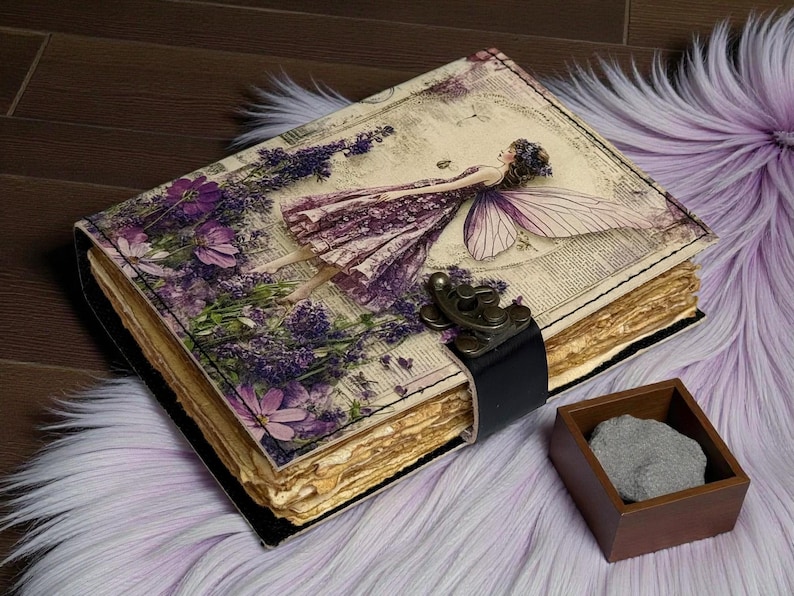 May include: A vintage-style journal with a fairy illustration on the cover. The cover features a purple fairy, lavender, and floral accents. The journal has a black leather strap closure and aged paper pages. A small wooden box with a gray stone sits nearby.