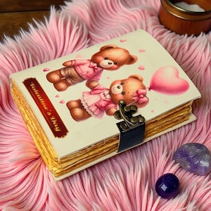 May include: A Valentine's Day journal with a teddy bear illustration on the cover. The journal has a metal clasp and is surrounded by a pink, fluffy surface. A pink heart-shaped balloon is also featured.